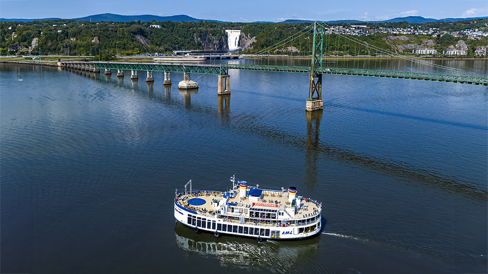 Un ferry blanc à plusieurs ponts navigue sur une large rivière, avec un pont suspendu vert visible au milieu de la scène. Au-delà du pont, une cascade dévale une falaise verdoyante. Un paysage vallonné et boisé borde la rivière, le tout sous un ciel bleu clair.