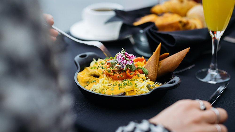 A colorful and elegant dish garnished with edible flowers, served in a small black skillet on a table with pastries, a cup of coffee, and a glass of juice.