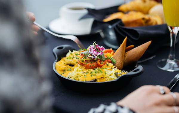 A colorful and elegant dish garnished with edible flowers, served in a small black skillet on a table with pastries, a cup of coffee, and a glass of juice.