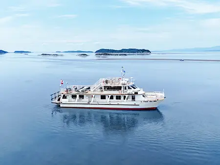 The ship "Vent des Îles" sails peacefully on calm waters surrounded by islands and a light mist on the horizon. The sky is clear and bright, creating a serene atmosphere.
