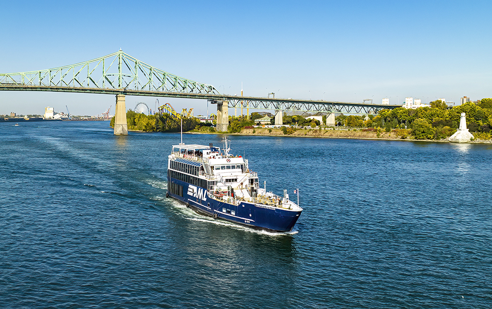 Un bateau de tourisme bleu navigue sur une large rivière sous un pont en acier vert. La scène ensoleillée révèle l'horizon d'une ville au loin et des arbres luxuriants bordant les rives, le tout sous un ciel bleu clair.