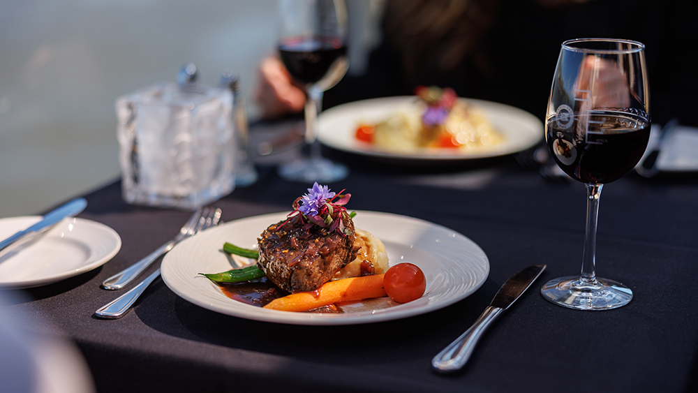 A beautifully plated dish with a piece of meat garnished with vegetables and edible flowers, accompanied by glasses of red wine on a table with a dark tablecloth.