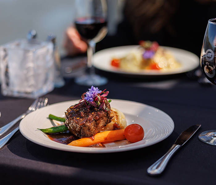 A beautifully plated dish with a piece of meat garnished with vegetables and edible flowers, accompanied by glasses of red wine on a table with a dark tablecloth.