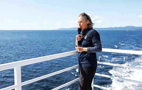 A naturalist stands on a boat's deck holding a microphone, overlooking the vast ocean under a clear blue sky. Distant landforms are visible across the water. The person wears a dark outfit and appears to be speaking or singing.