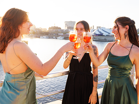 Three women dressed in elegant attire toasting with cocktails on the terrace of the AML Cavalier Maxim ship in Montreal