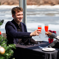 A smiling and affectionate couple clinking glasses with a cocktail at a table during a Christmas brunch cruise, with a decorated Christmas tree in the foreground on the left