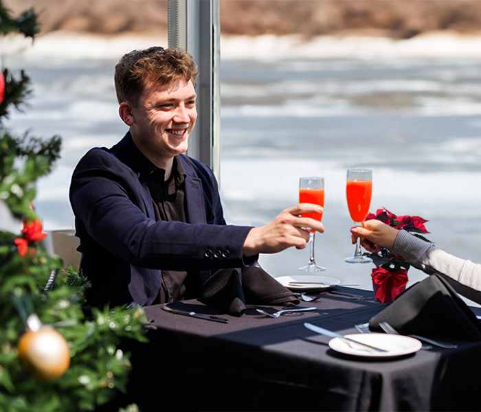 A smiling and affectionate couple clinking glasses with a cocktail at a table during a Christmas brunch cruise, with a decorated Christmas tree in the foreground on the left