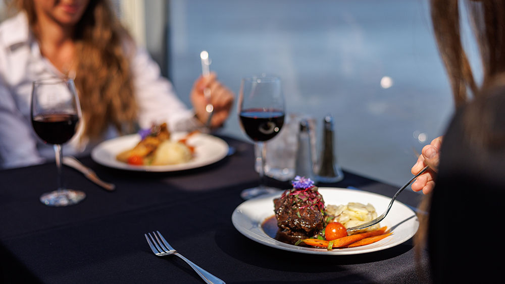 Two people enjoying a gourmet meal on an elegant table with a black tablecloth, including red wine and beautifully plated dishes, with a view of the water in the background.