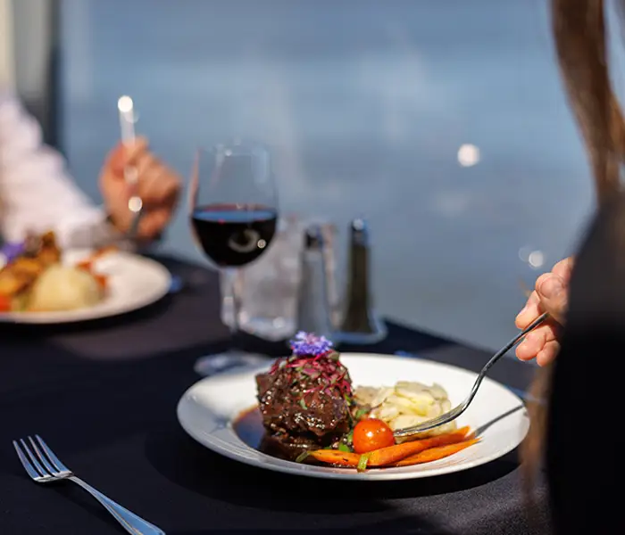 Deux personnes dégustent un repas gastronomique sur une table élégante avec une nappe noire, comprenant du vin rouge et des plats soigneusement présentés, le tout avec une vue sur l'eau en arrière-plan.