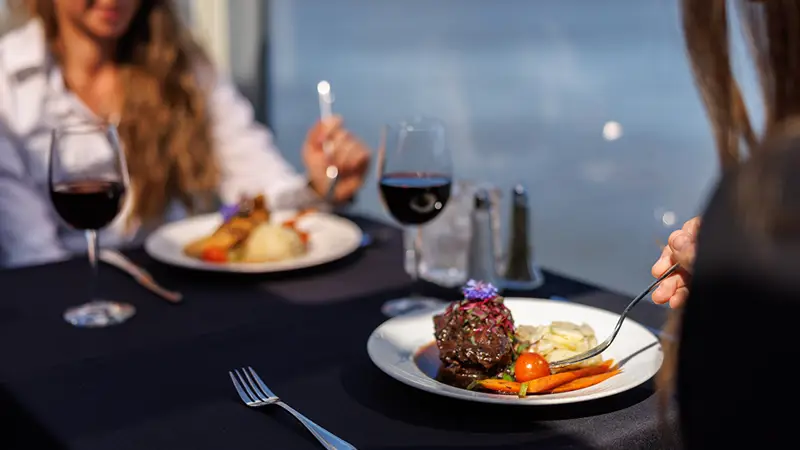 Deux personnes dégustent un repas gastronomique sur une table élégante avec une nappe noire, comprenant du vin rouge et des plats soigneusement présentés, le tout avec une vue sur l'eau en arrière-plan.
