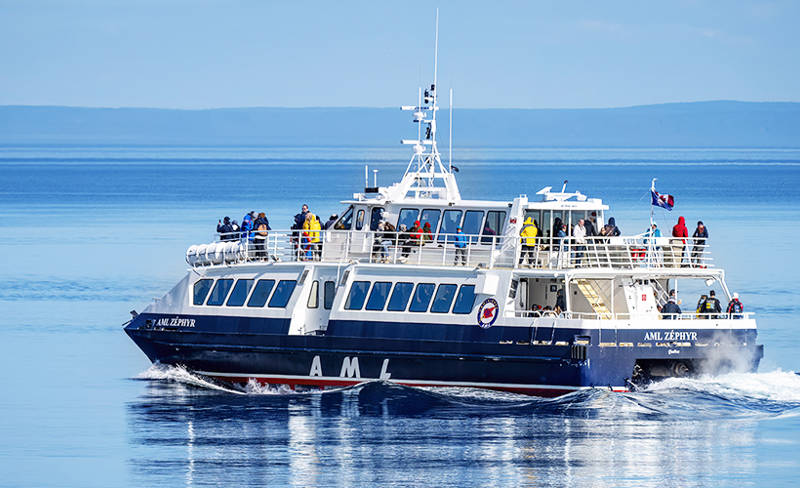 The ship "AML Zéphyr" moves across calm waters, carrying passengers enjoying a beautiful day. The boat, blue with white accents, leaves a light trail in the water. Distant hills are visible on the horizon under a clear sky.