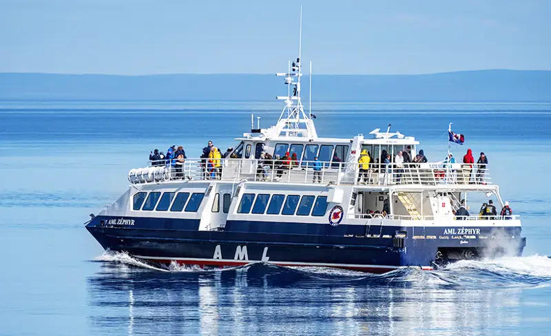 The ship "AML Zéphyr" moves across calm waters, carrying passengers enjoying a beautiful day. The boat, blue with white accents, leaves a light trail in the water. Distant hills are visible on the horizon under a clear sky.