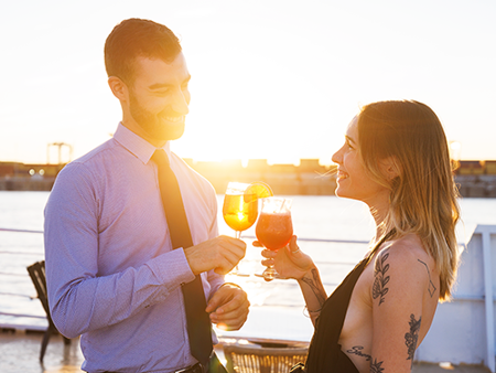 A smiling and affectionate couple enjoying a cocktail on the terrace of the AML Cavalier Maxim ship in Montreal, with a sunset in the background