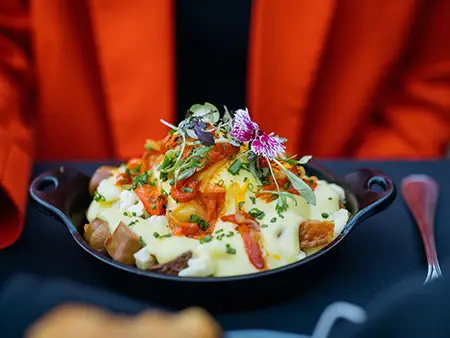 A delicious dish of roasted potatoes topped with hollandaise sauce, garnished with vegetables, fresh herbs, and edible flowers, served in a cast-iron skillet.