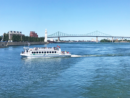 A river shuttle crosses the water under a blue sky, with the Clock Tower, Jacques Cartier Bridge, and Montreal buildings visible in the background.