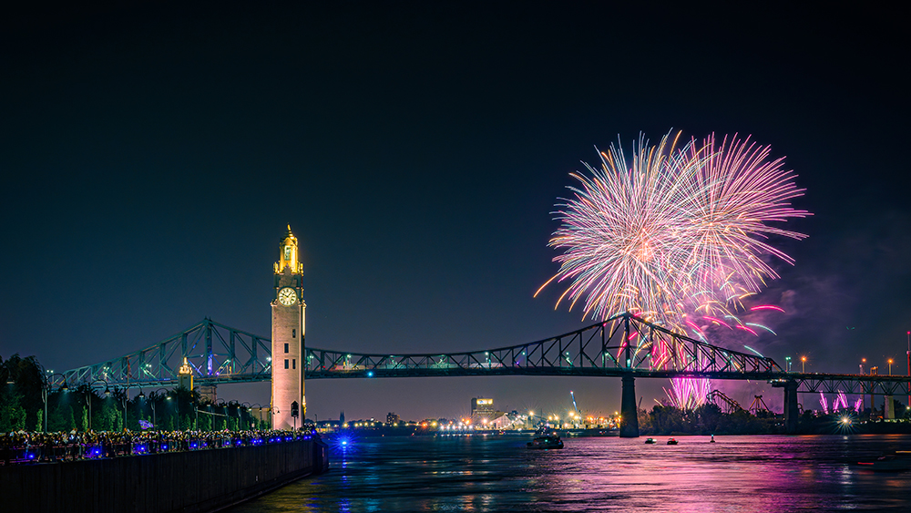 Colorful fireworks burst in the night sky near the Jacques Cartier Bridge and the Clock Tower, with reflections on the river and a crowd watching from the quay.