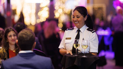 A smiling waitress in uniform serves two glasses of white wine to guests in an elegant setting with dim lights and a festive atmosphere.