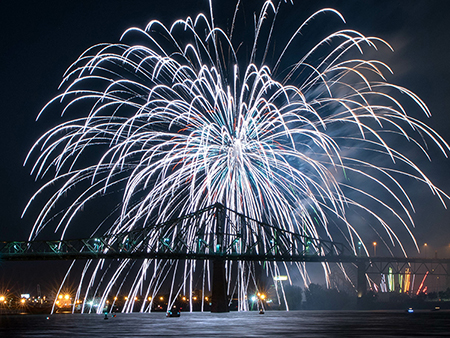Un feu d'artifice spectaculaire éclate dans le ciel nocturne au-dessus d'un pont illuminé, avec des reflets sur l'eau et des lumières urbaines en arrière-plan.