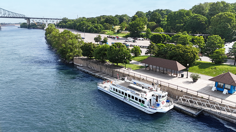 Une vue aérienne d'un bateau de croisière amarré le long d'un quai bordé d'arbres, avec un parc verdoyant en arrière-plan et un pont visible au loin.
