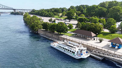 An aerial view of a cruise boat docked along a waterfront lined with trees, with a green park in the background and a bridge visible in the distance.