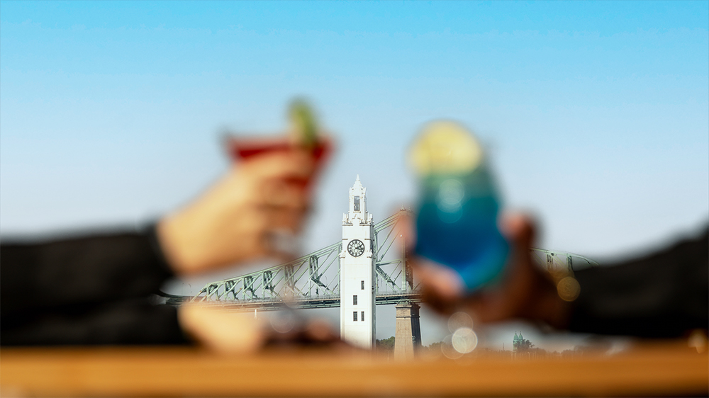 Two people toasting with colorful cocktails (one red and one blue) against a blurred background of a white clock tower and a bridge.
