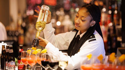 A bartender in uniform prepares cocktails by pouring liquor into glasses garnished with orange slices, in a well-lit bar with bottles in the background.