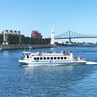 Une navette fluviale traverse le fleuve sous un ciel bleu, avec la tour de l'Horloge, le pont Jacques-Cartier et des bâtiments de Montréal visibles en arrière-plan.