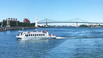 Une navette fluviale traverse le fleuve sous un ciel bleu, avec la tour de l'Horloge, le pont Jacques-Cartier et des bâtiments de Montréal visibles en arrière-plan.