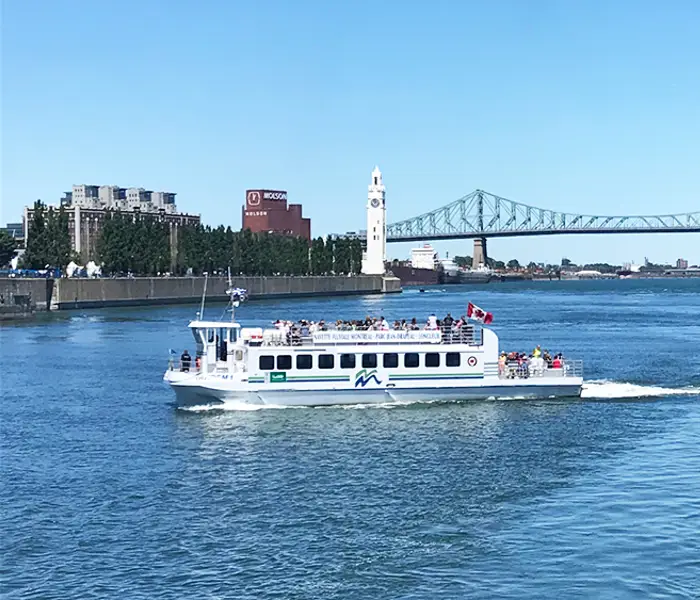 Une navette fluviale traverse le fleuve sous un ciel bleu, avec la tour de l'Horloge, le pont Jacques-Cartier et des bâtiments de Montréal visibles en arrière-plan.