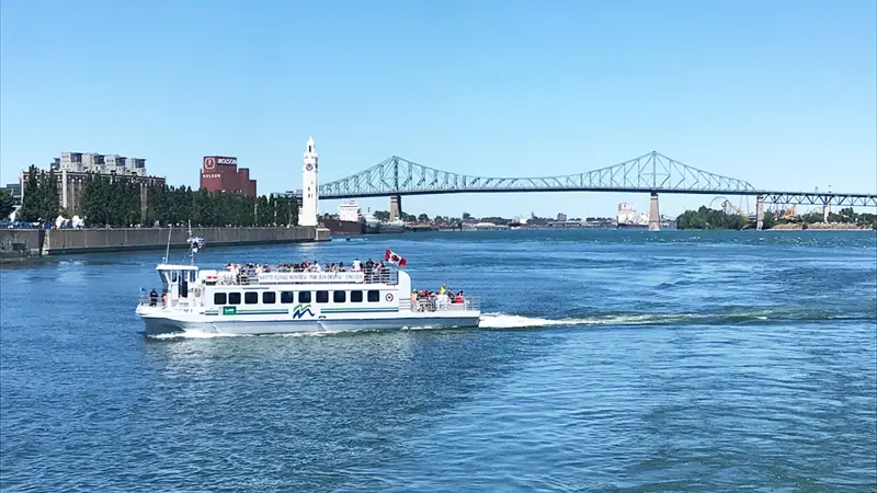 Une navette fluviale traverse le fleuve sous un ciel bleu, avec la tour de l'Horloge, le pont Jacques-Cartier et des bâtiments de Montréal visibles en arrière-plan.