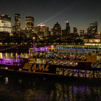  Un bateau de croisière "AML" illuminé navigue sur une rivière la nuit, avec une vue spectaculaire sur les gratte-ciels et les lumières de la ville en arrière-plan.