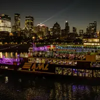 An illuminated "AML" cruise ship sails on a river at night, with a stunning view of skyscrapers and city lights in the background.