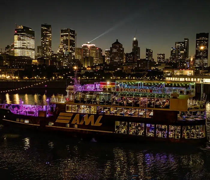 An illuminated "AML" cruise ship sails on a river at night, with a stunning view of skyscrapers and city lights in the background.