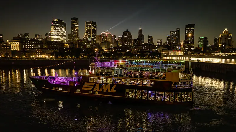 An illuminated "AML" cruise ship sails on a river at night, with a stunning view of skyscrapers and city lights in the background.