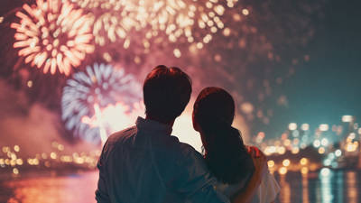 A couple watches a colorful fireworks display lighting up the night sky, with shimmering reflections on the water and city lights in the background.