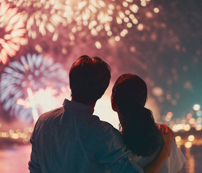 A couple watches a colorful fireworks display lighting up the night sky, with shimmering reflections on the water and city lights in the background.