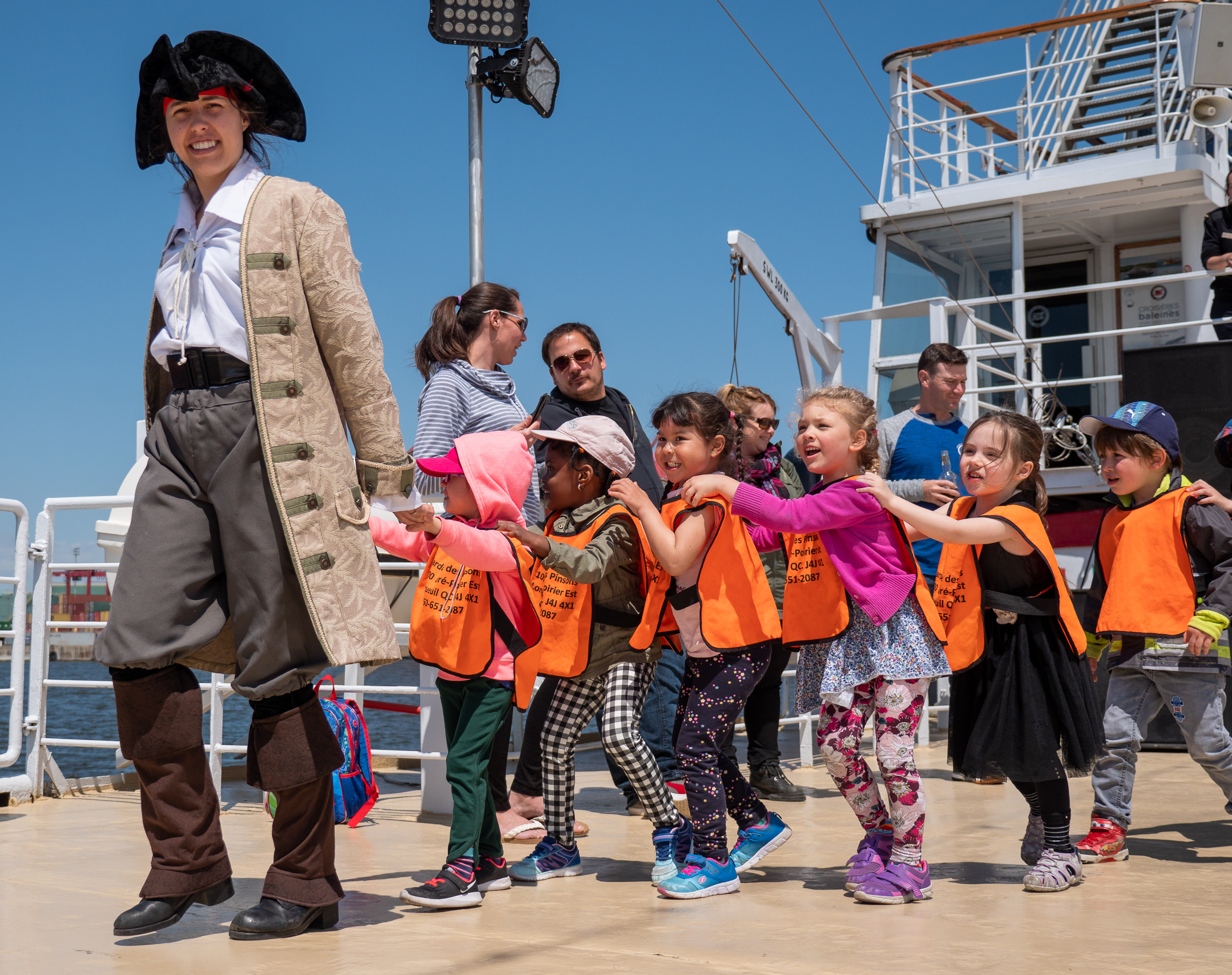 A person dressed as a pirate leads a line of children wearing orange safety vests on a ship deck. The kids are smiling, holding onto each other, and enjoying the experience. In the background, adults observe and a clear blue sky is visible.