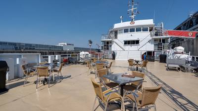 One of the outdoor terraces of the AML Cavalier Maxim ship under a blue sky. Tables and chairs are set up, and the ship is docked.