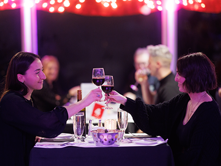 Couple souriant et complice assis à une table trinquant avec un verre de vin rouge avec des décorations de Noël en arrière plan