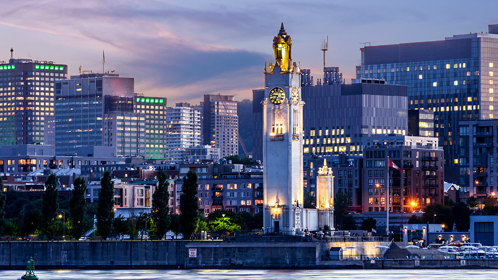 The Clock Tower rises by the river, lit up at dusk, with buildings and skyscrapers illuminated in the background under a twilight sky.