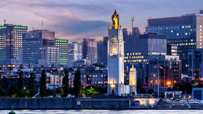 The Clock Tower rises by the river, lit up at dusk, with buildings and skyscrapers illuminated in the background under a twilight sky.