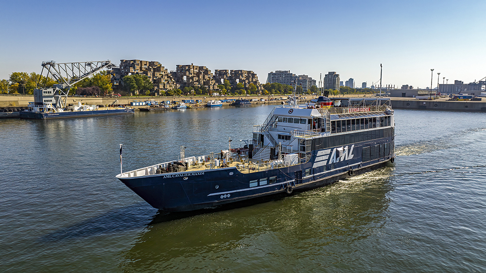 A black cruise ship with "AML" written on it, sailing on a river. In the background, a distinctive residential complex, a bridge, and urban buildings under a clear blue sky.