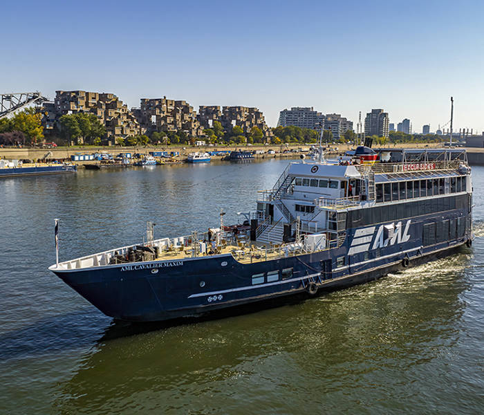 A black cruise ship with "AML" written on it, sailing on a river. In the background, a distinctive residential complex, a bridge, and urban buildings under a clear blue sky.