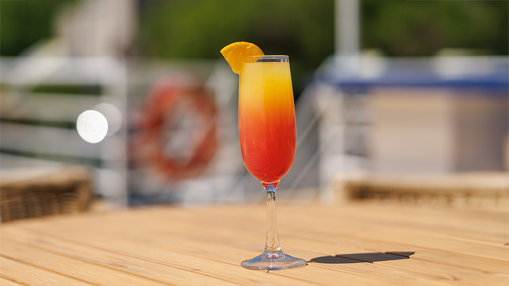 A colorful red and yellow cocktail glass garnished with an orange slice, placed on a wooden table with a blurred background featuring a boat and nautical elements.