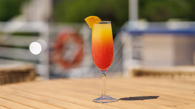 A colorful red and yellow cocktail glass garnished with an orange slice, placed on a wooden table with a blurred background featuring a boat and nautical elements.