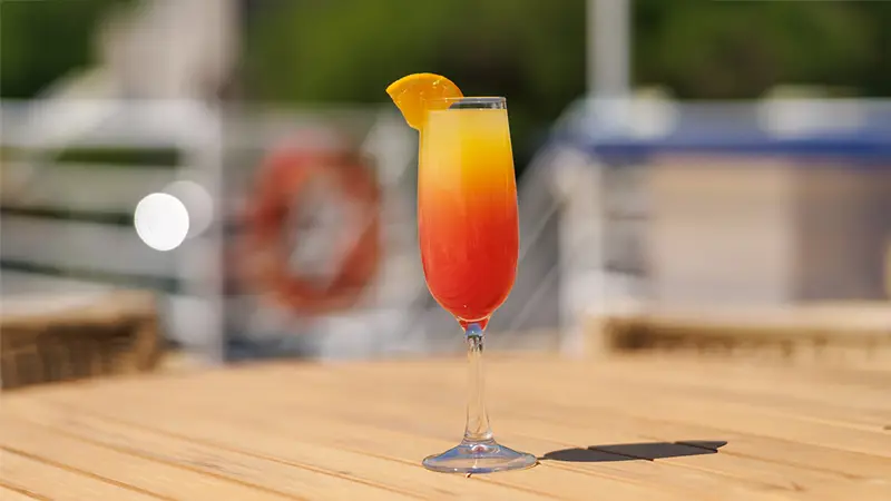 A colorful red and yellow cocktail glass garnished with an orange slice, placed on a wooden table with a blurred background featuring a boat and nautical elements.