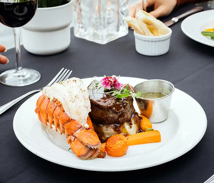 A gourmet dish featuring filet mignon, a lobster tail, grilled vegetables, and a side of sauce, served on an elegant table with a black tablecloth. A glass of red wine and another dish are also visible nearby.