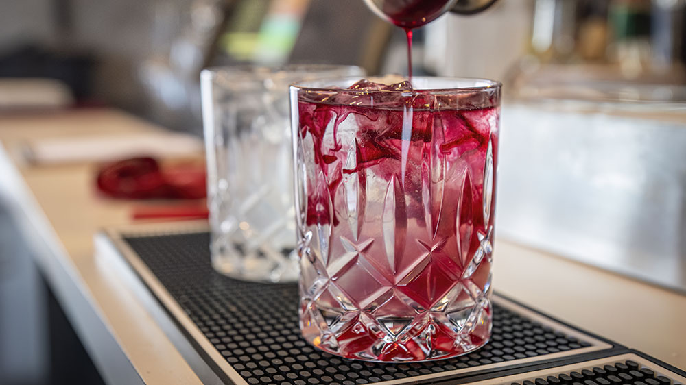 A cocktail is being prepared in a clear crystal glass filled with ice cubes. Red liquid is being poured, creating a swirling effect with the ice. In the background, another similar glass and blurred bar elements are visible.