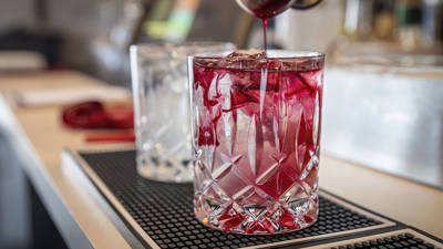 A cocktail is being prepared in a clear crystal glass filled with ice cubes. Red liquid is being poured, creating a swirling effect with the ice. In the background, another similar glass and blurred bar elements are visible.