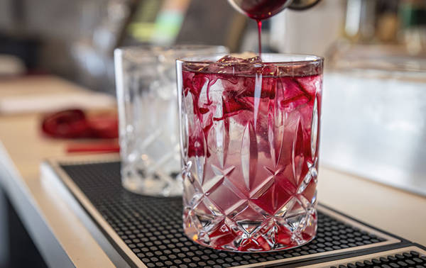 A cocktail is being prepared in a clear crystal glass filled with ice cubes. Red liquid is being poured, creating a swirling effect with the ice. In the background, another similar glass and blurred bar elements are visible.